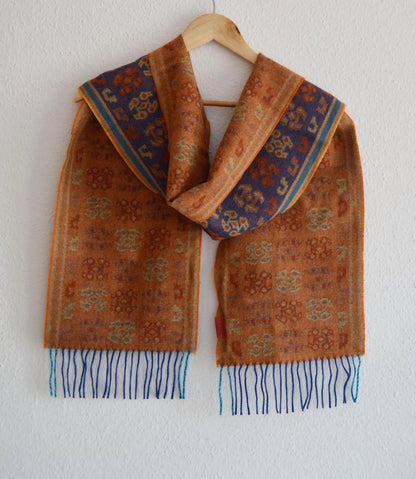 Handmade brown and blue alpaca shawl with intricate patterns and fringed edges hanging on wooden hanger.