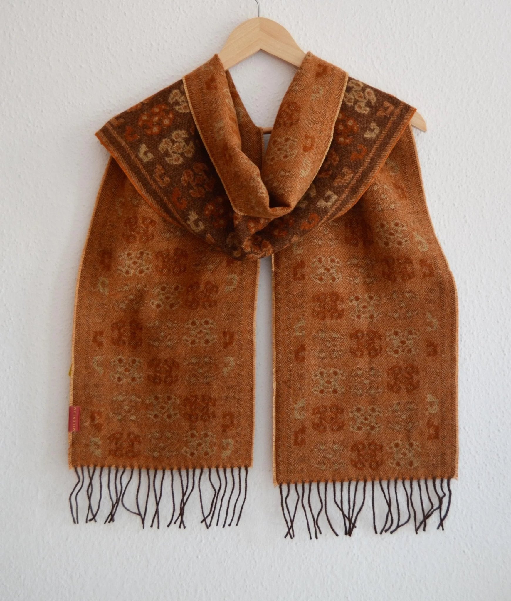 Handmade brown alpaca shawl with traditional patterns and fringe edges hanging on wooden hanger.
