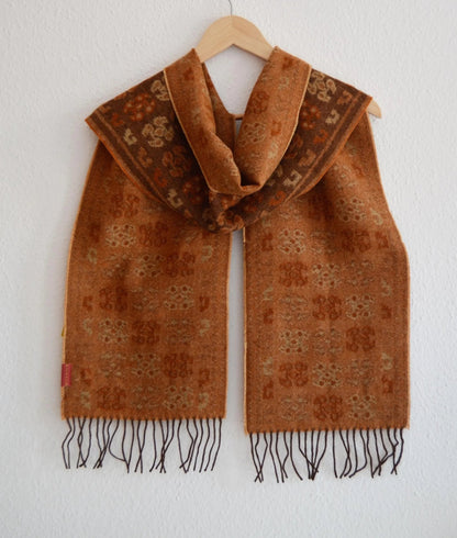 Handmade brown alpaca shawl with traditional patterns and fringe edges hanging on wooden hanger.