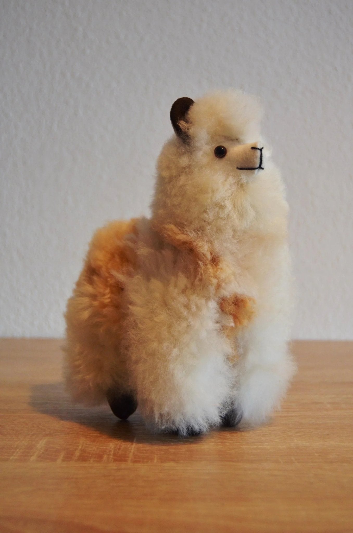 Handmade soft alpaca plush in cream and light brown with black eyes and nose, standing on wood surface.