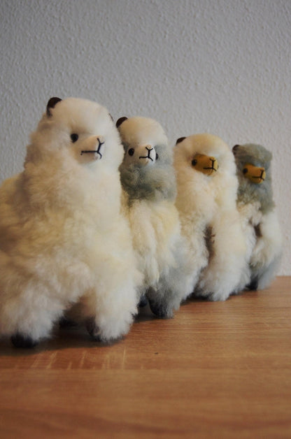 Four handmade alpaca plush toys in white, white-gray, gray, and brown with soft wool texture, standing on wood surface.